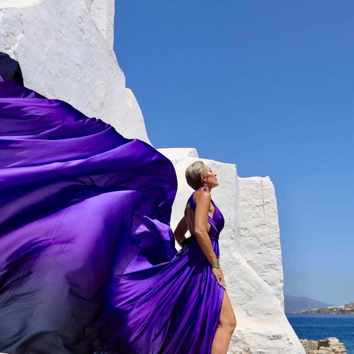 Woman in flowing purple dress against white structures and clear blue sky.