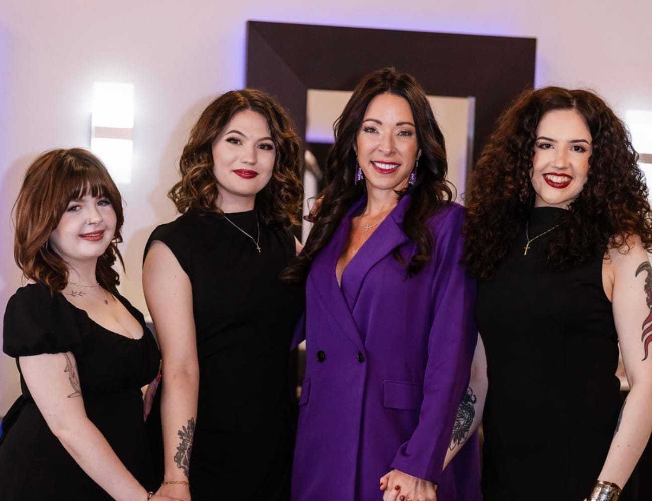 Four women smiling, one in purple, the others in black dresses, posing indoors.